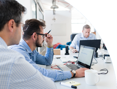 Eine Runde von Personen am Tisch, alle mit Laptops, die gemeinsam arbeiten. Die Person in der Mitte wirkt nachdenklich und hält einen Stift in der linken Hand.